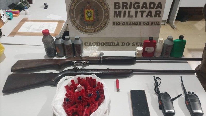 Sobre uma mesa est&aacute; uma espingarda calibre .28, um rifle calibre .22, um cartucho calibre .28, 83 cartuchos deflagrados, dois r&aacute;dios comunicadores, sete potes de p&oacute;lvora, um pote contendo chumbo, um saco pl&aacute;stico contendo chumbo e um aparelho celular.