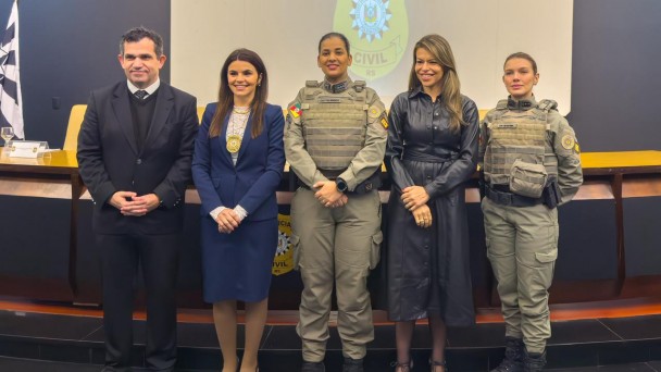 Imagem mostra cinco pessoas posando em um audit&oacute;rio, em frente a uma mesa e a uma tela com o bras&atilde;o da Pol&iacute;cia Civil projetado ao fundo. Duas policiais militares aparecem fardadas, ao centro e &agrave; direita da imagem, e tr&ecirc;s pessoas est&atilde;o em trajes civis. Todos est&atilde;o em p&eacute;, lado a lado, voltados para a c&acirc;mera.